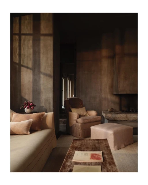 Living room interior features a sofa with pillows, an armchair, an ottoman, a coffee table with books, and window light on the wall.