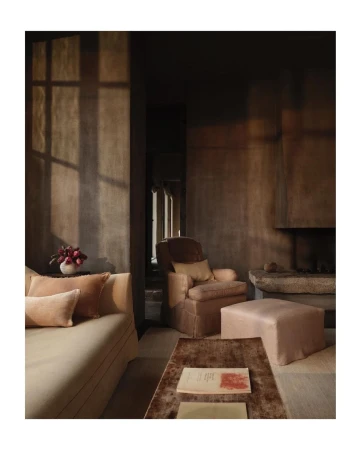 Living room interior features a sofa with pillows, an armchair, an ottoman, a coffee table with books, and window light on the wall.