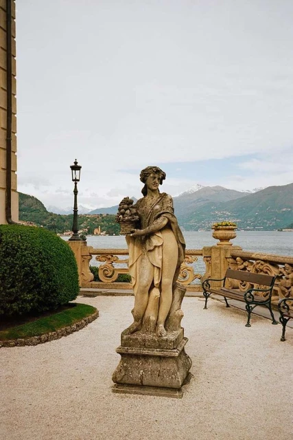 Stone statue of woman on a terrace overlooking Lake Como, Italy, with mountains in the background, ornate railing and bench.
