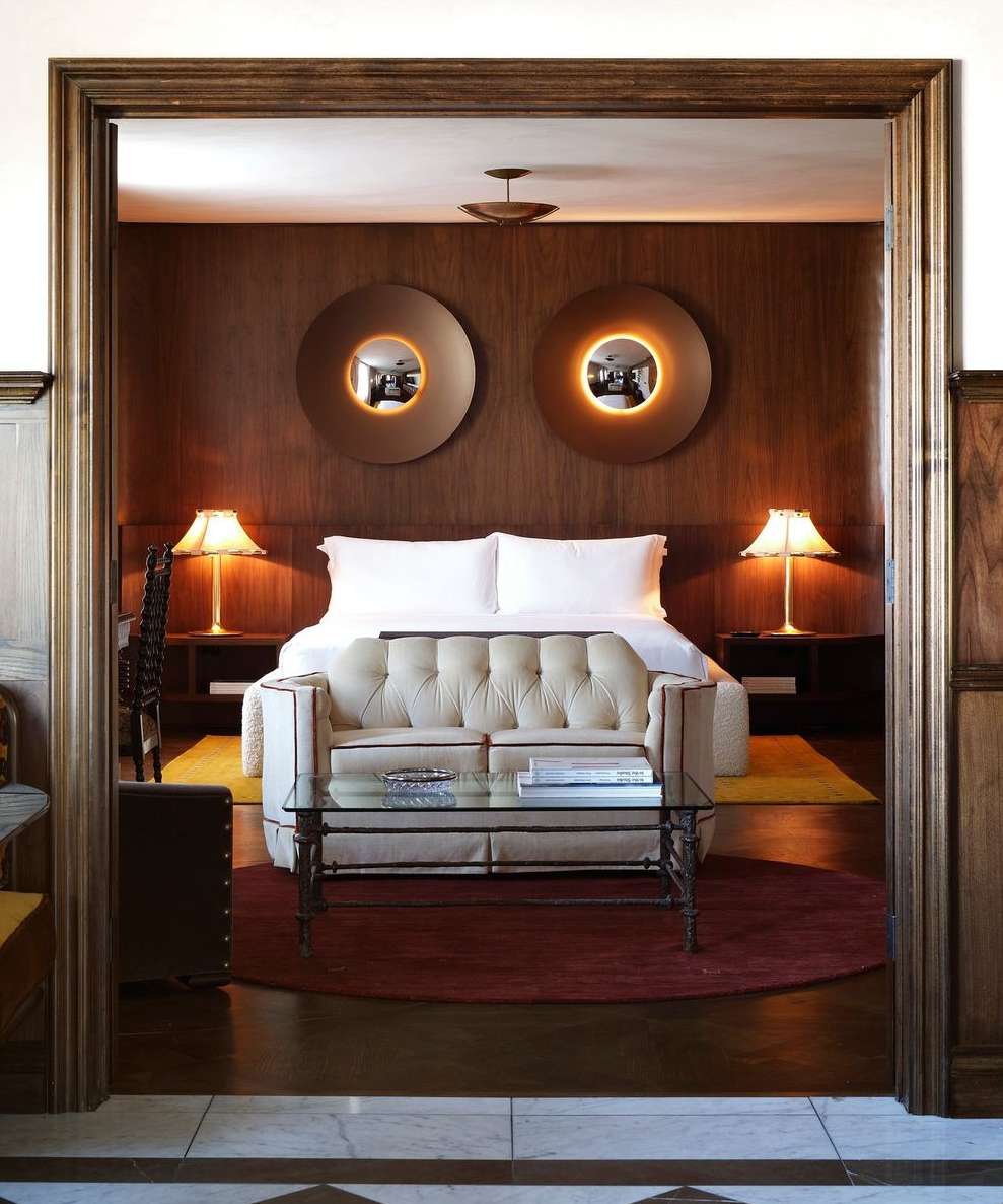 Hotel room interior featuring a tufted sofa, glass coffee table, bed with white linens, and round mirrors on a dark wood paneled wall.