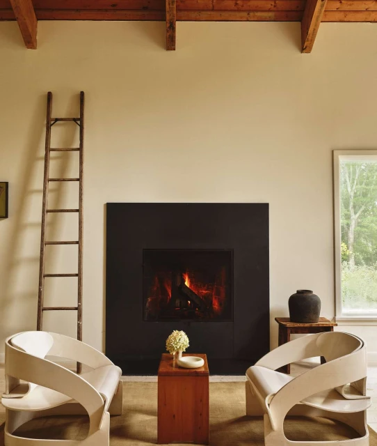 Interior of a living room featuring a fireplace with a black frame, two armchairs, a ladder, and a wooden coffee table.