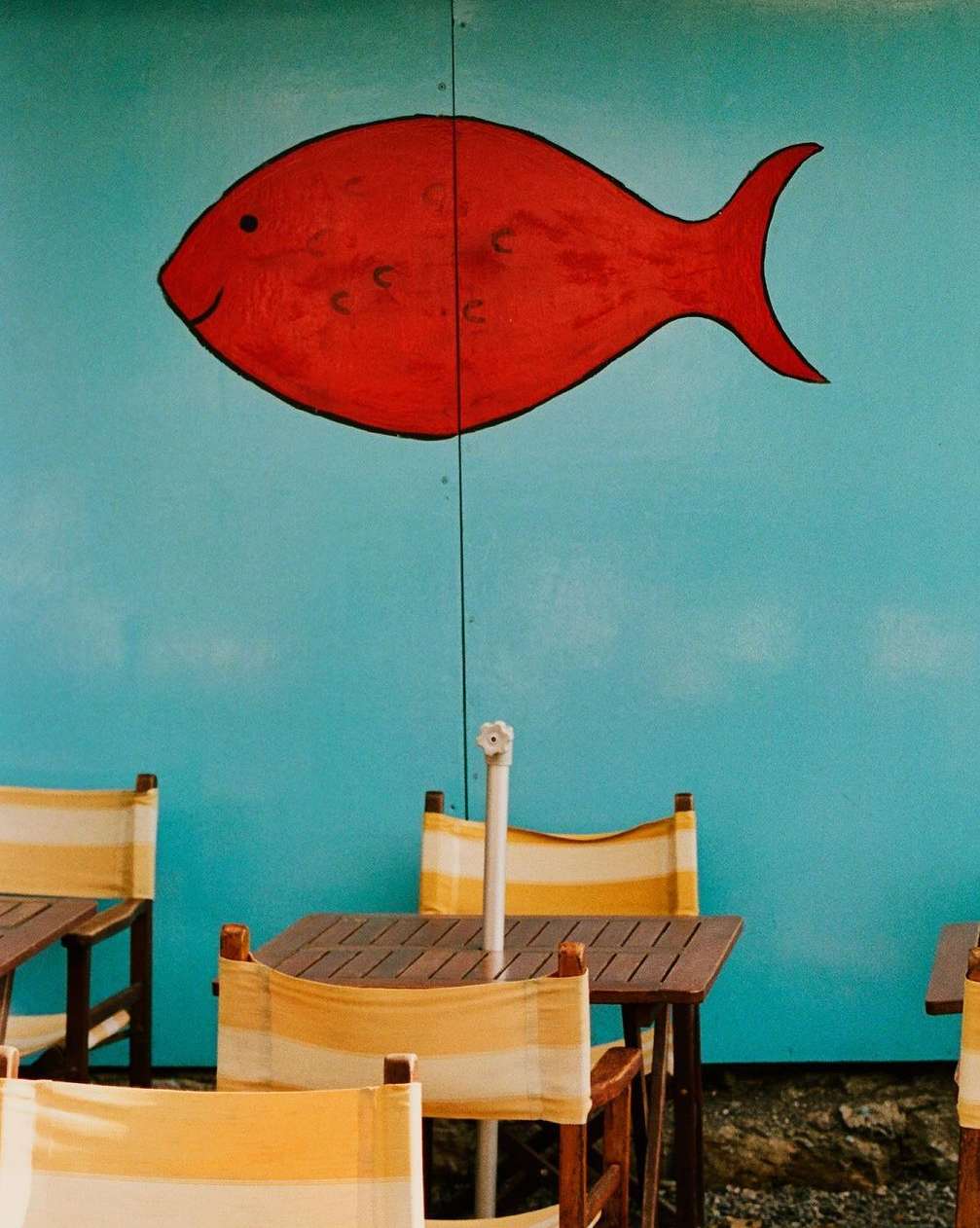 Outdoor cafe featuring striped seating and a red fish mural on a turquoise wall, creating a vibrant, summery scene.