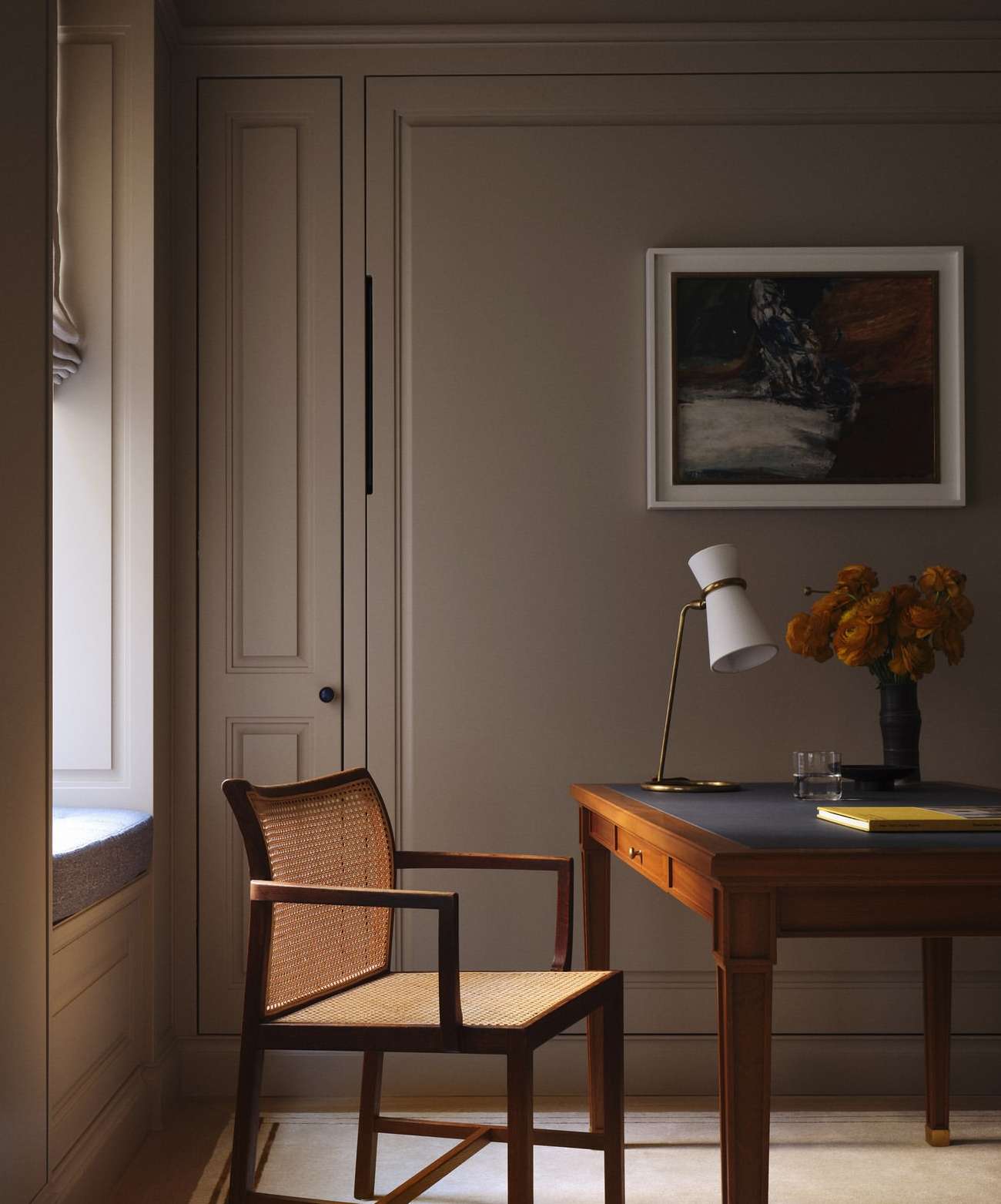 Home office interior with a cane chair, wooden desk, table lamp, and a framed painting, illuminated by natural light from a nearby window.