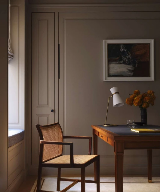 Home office interior with a cane chair, wooden desk, table lamp, and a framed painting, illuminated by natural light from a nearby window.