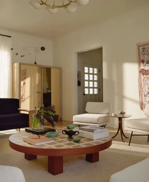 Living room interior with a round wood and tile coffee table, sofa, armchairs, and decorative wall hanging in soft, natural light.