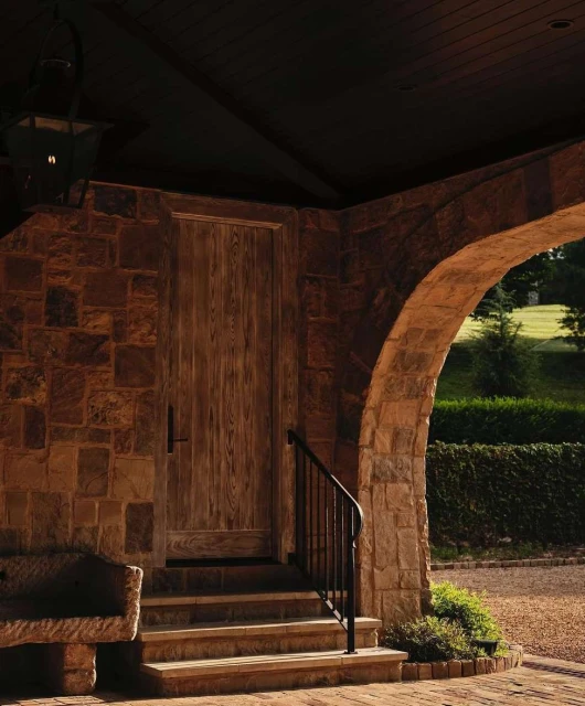 Stone archway leads to a wooden door with black hardware, steps, and landscaping in a residential setting under natural light.