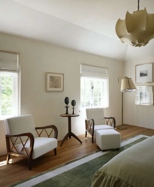 Well-lit bedroom interior featuring two white armchairs, a round table with decor, a floor lamp, and a striped green rug over wood flooring.