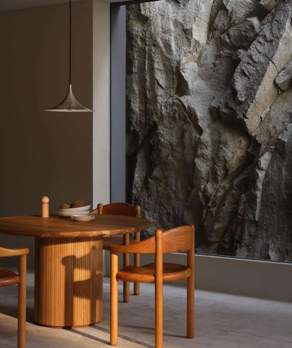 A round, light-colored wood dining table and chairs set in an interior with a rock feature wall by a window and modern pendant light.