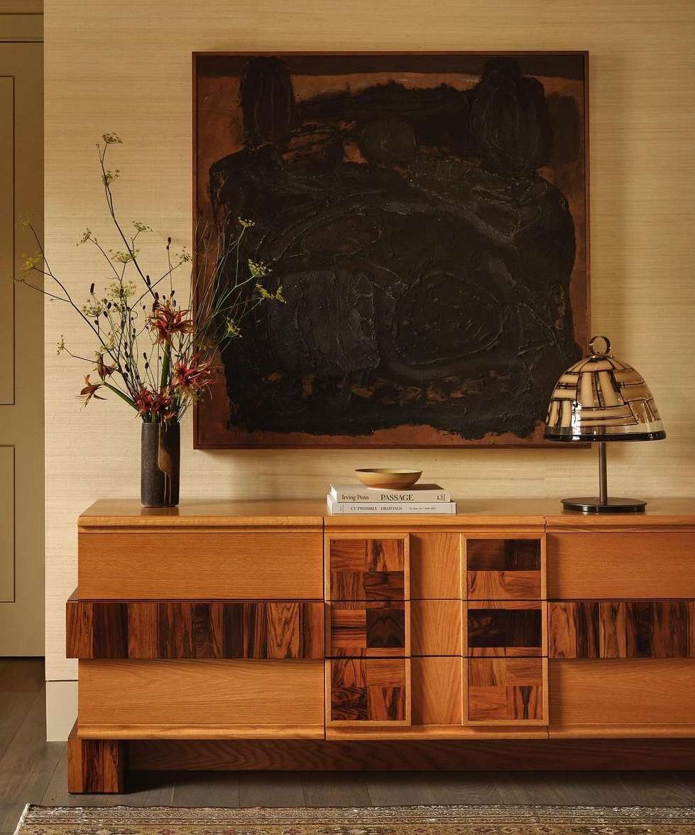 Interior shot of a credenza beneath an abstract painting, featuring a lamp, flowers in a vase, and books, set against a patterned wall.