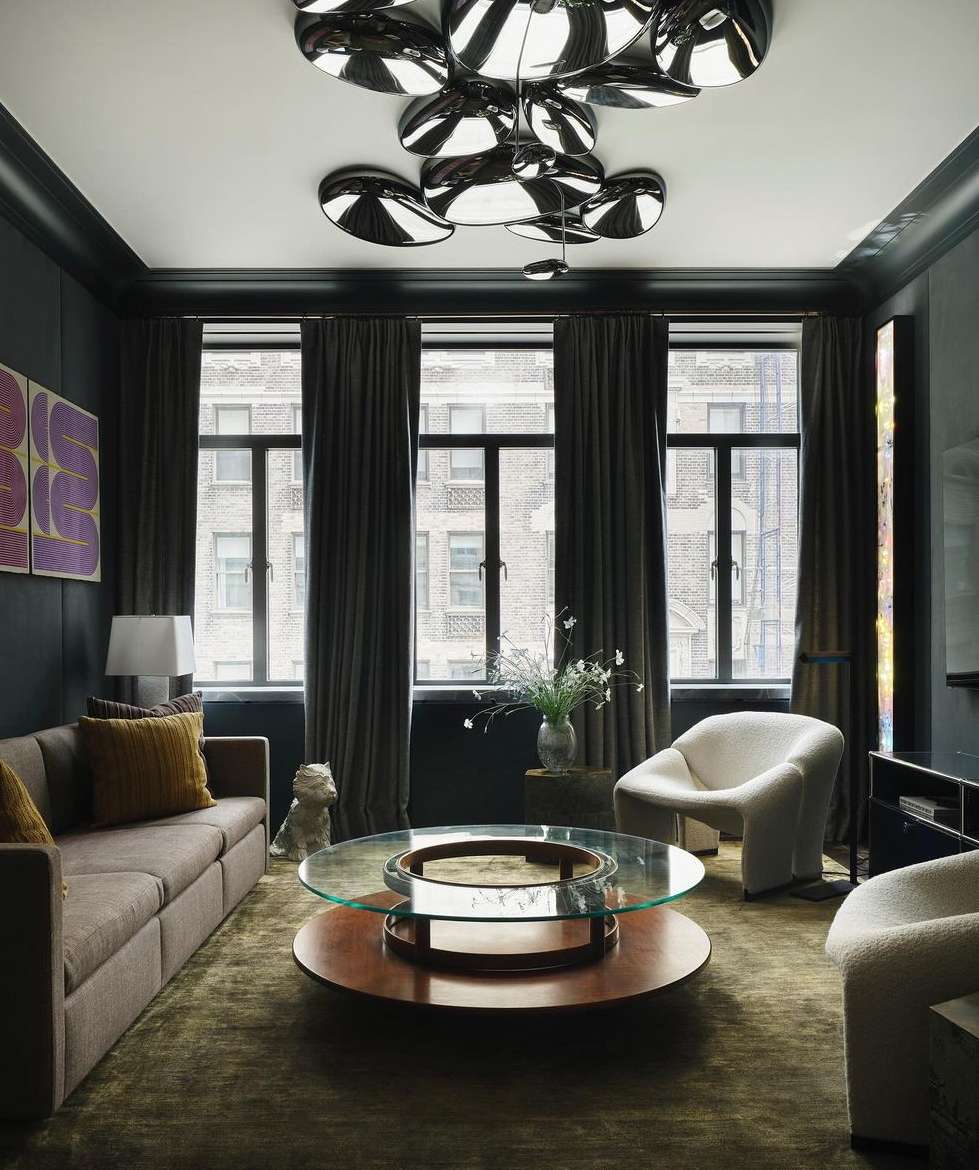 Living room interior featuring a brown couch, round coffee table, and armchairs, illuminated by modern disc lighting. Dark walls and window.