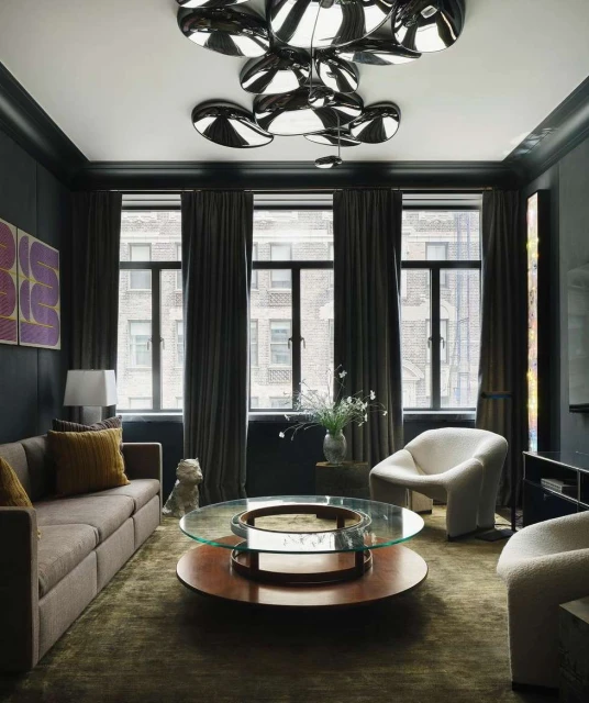 Living room interior featuring a brown couch, round coffee table, and armchairs, illuminated by modern disc lighting. Dark walls and window.