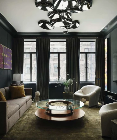 Living room interior featuring a brown couch, round coffee table, and armchairs, illuminated by modern disc lighting. Dark walls and window.