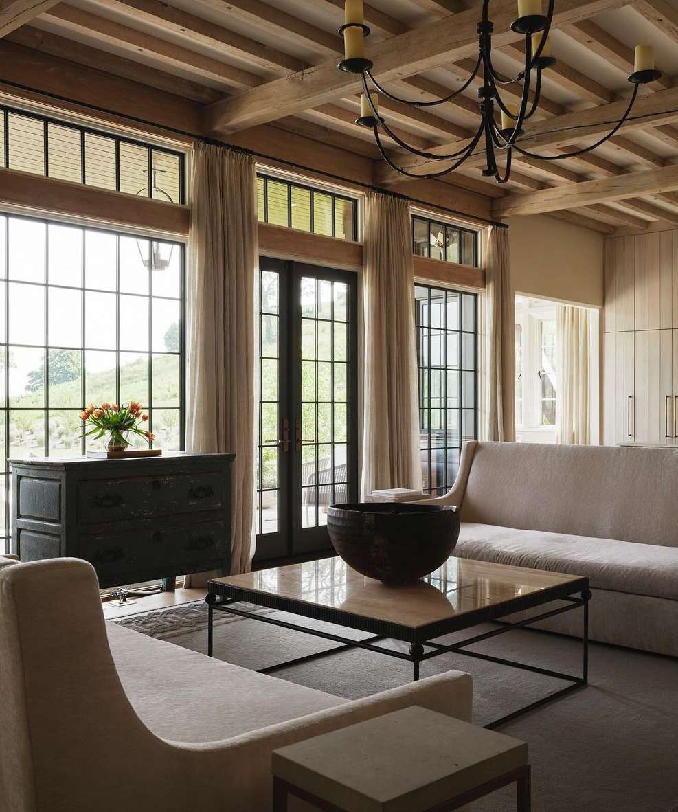 Living room interior with neutral sofas, black console table, coffered wood ceiling, large windows, natural light, and indoor plants.