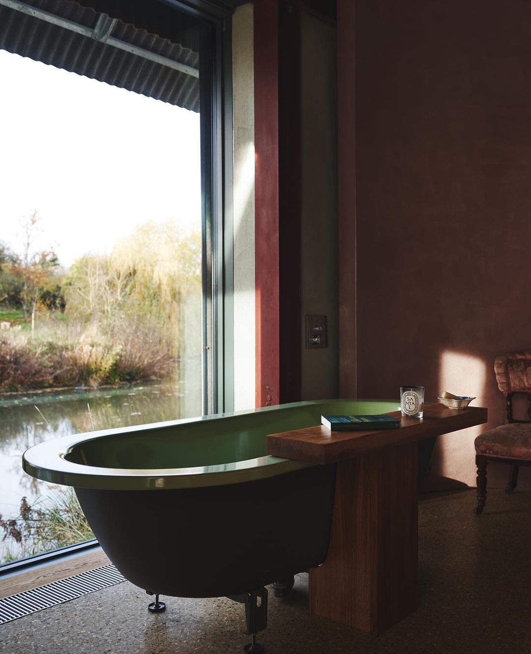 Olive-green bathtub in a modern bathroom with a large window overlooking a pond and trees, with muted tones and natural light.