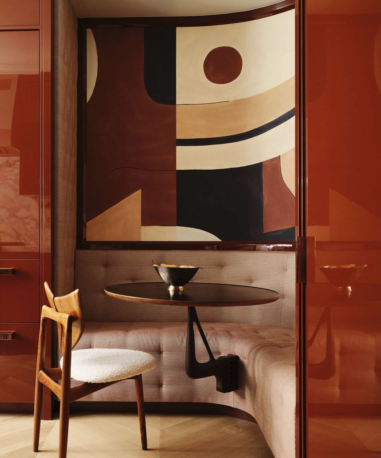 Interior featuring a wood chair and table next to a cushioned bench, with an abstract painting on the wall and an orange cabinet.