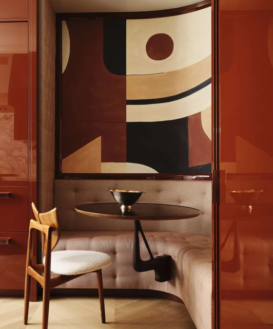 Interior featuring a wood chair and table next to a cushioned bench, with an abstract painting on the wall and an orange cabinet.