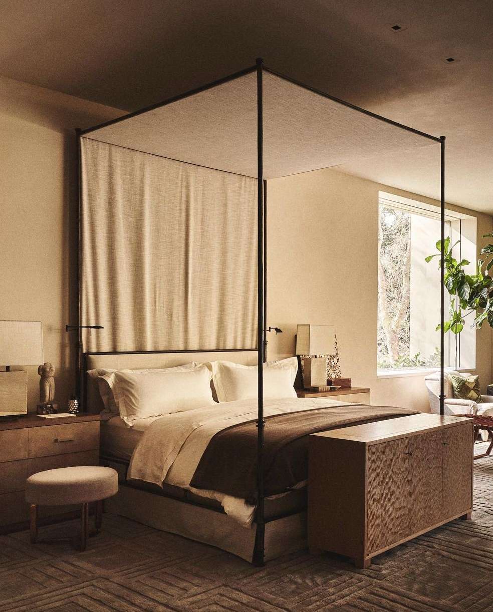 Well-lit bedroom interior features a four-poster bed with neutral-toned bedding, side tables, a stool, and a carpet with a geometric pattern.