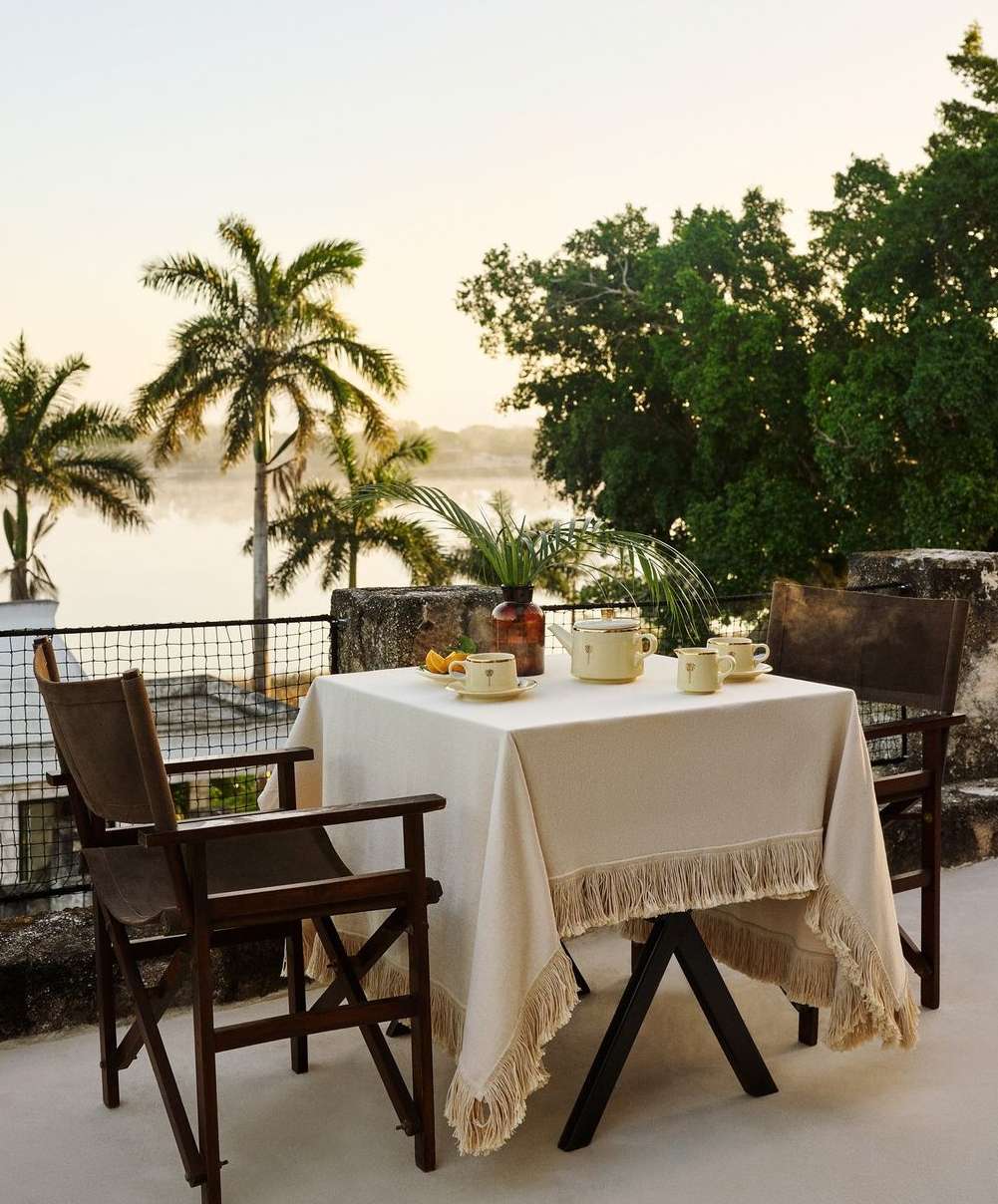 Outdoor dining table with fringed linen, set with teacups and a teapot, flanked by two wooden chairs on a stone patio.