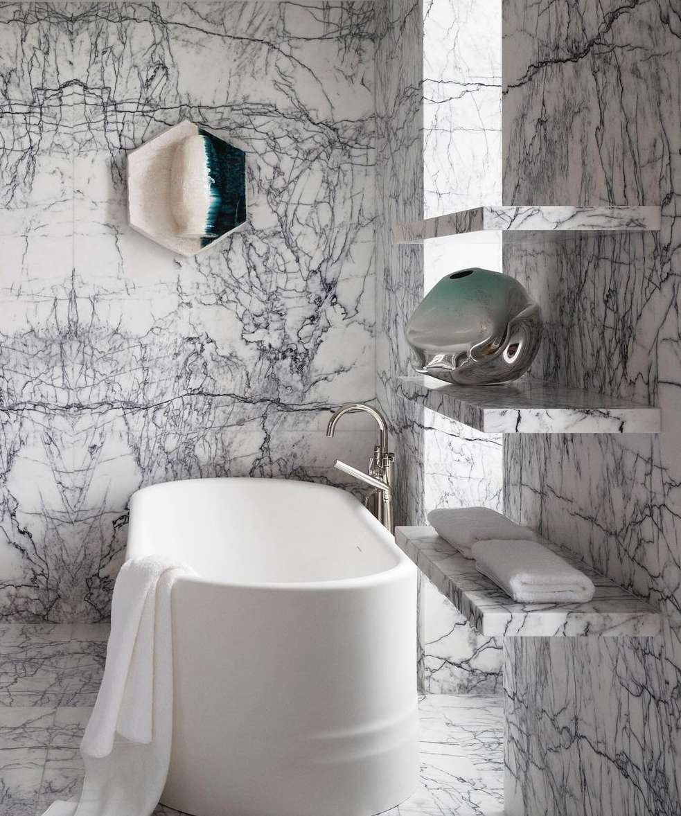 Modern bathroom interior featuring a white bathtub, marble walls and shelves, and decorative vases in a bright, clean setting.