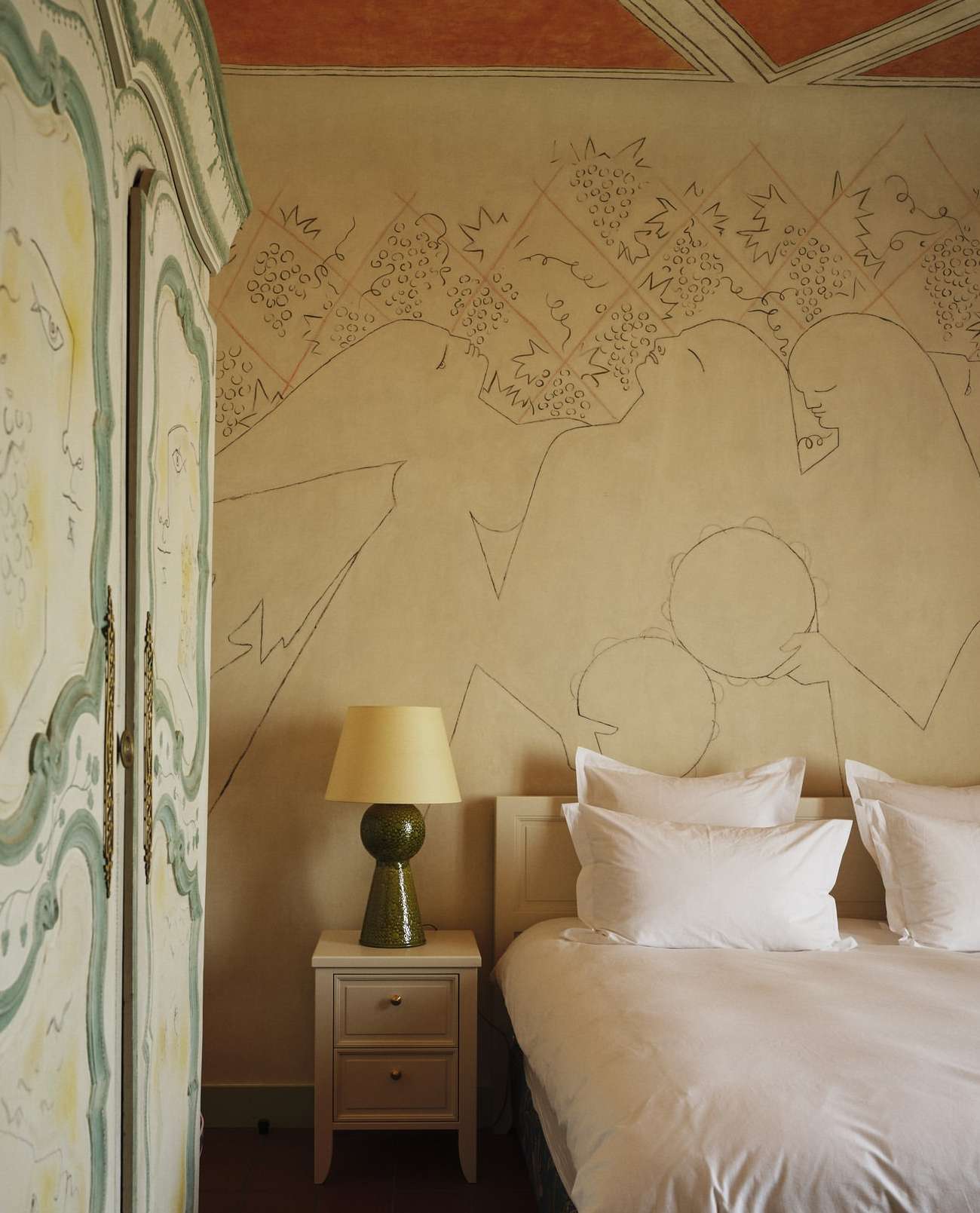 Bedroom interior featuring a bed with white linens, a bedside table with a lamp, and an ornate wardrobe, all set against a wall mural.