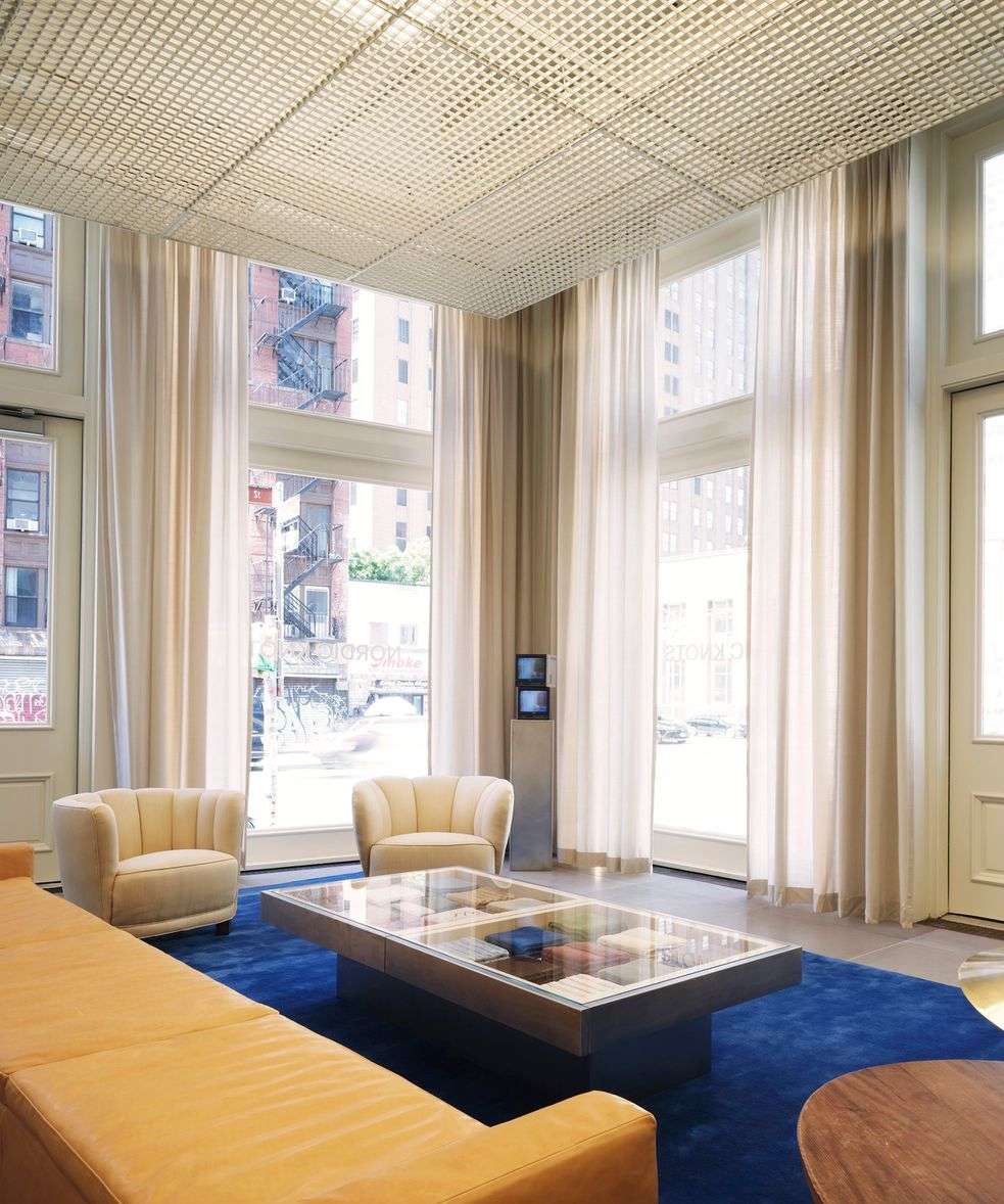 Living room interior with a long tan leather sofa, two cream-colored armchairs, a blue rug, and tall windows with sheer white curtains.