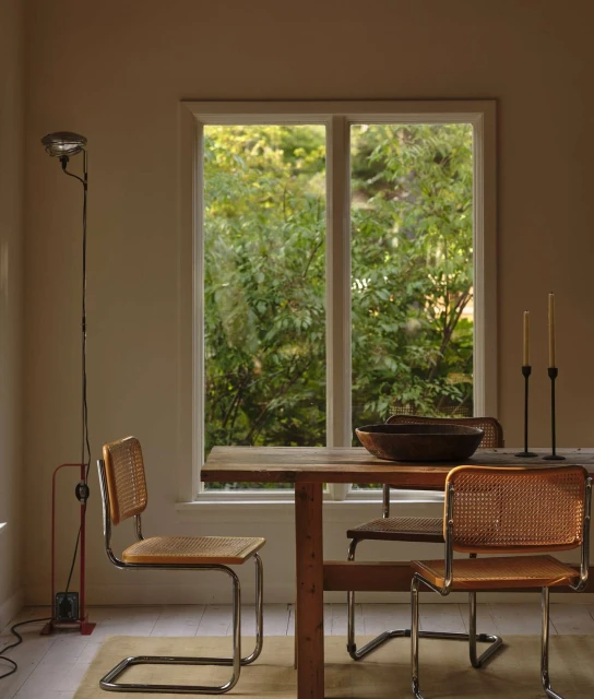 Interior view of a rustic wooden dining table with four cane chairs near a bright window, lit by a standing lamp and candles.