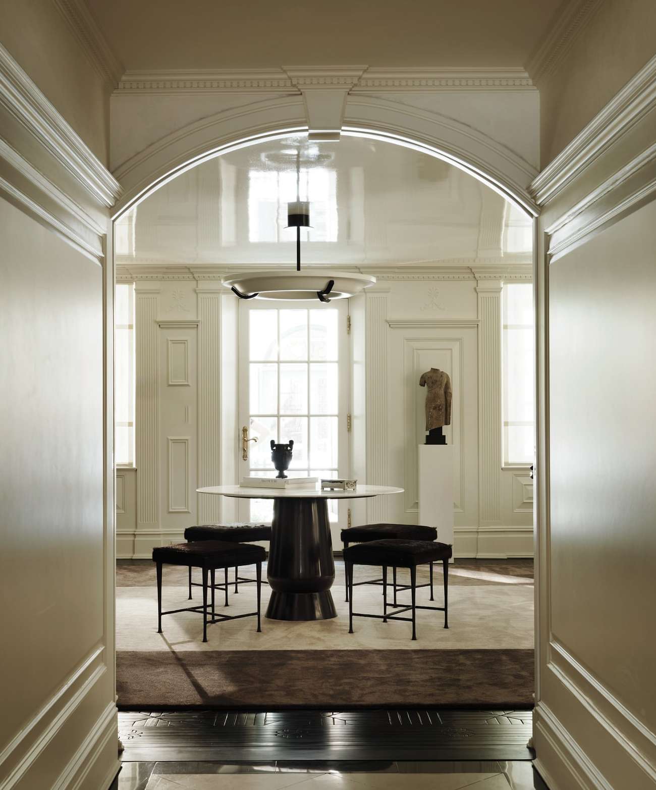 Elegant interior features a round marble-top table with dark base surrounded by four square stools, viewed through an arched doorway.