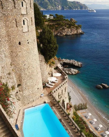 Scenic view of a stone building with a swimming pool overlooking a beach and the ocean with mountains in the background.