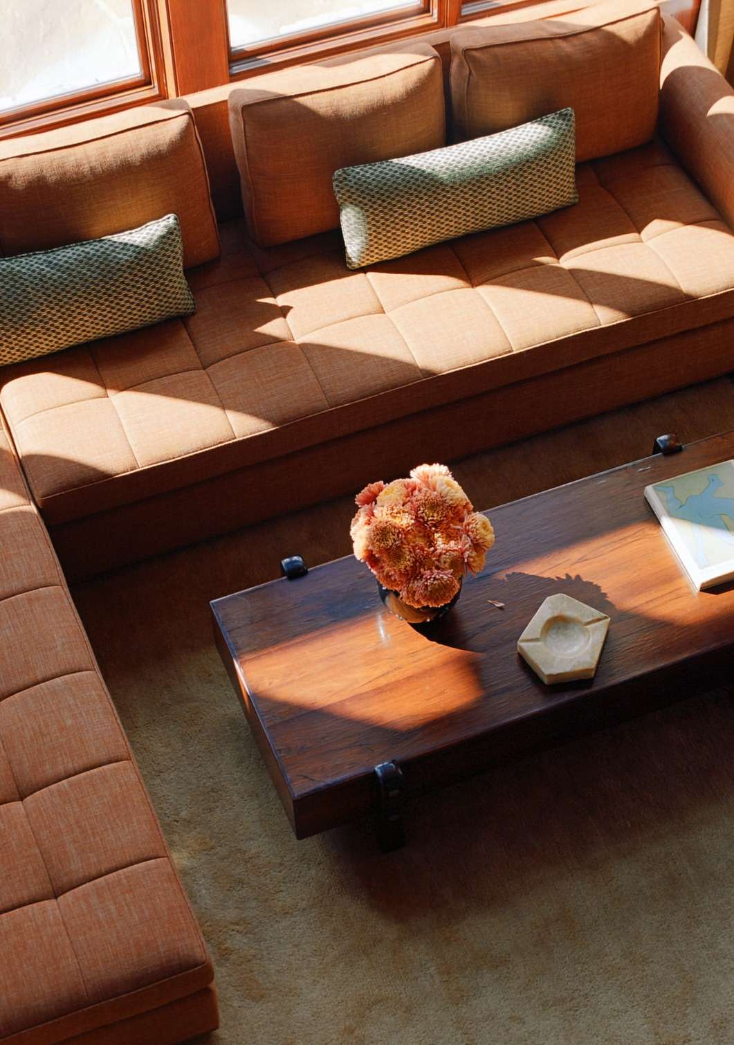 Modern interior featuring a tan sectional sofa, dark wood coffee table with floral arrangement, light-colored rug, and natural light.