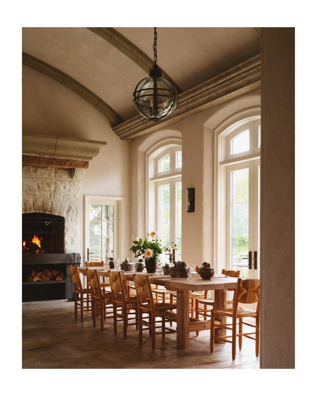 Interior view of a dining room with a long wooden table, chairs, stone fireplace with burning logs, and natural light from large windows.