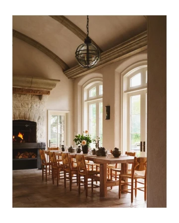 Interior view of a dining room with a long wooden table, chairs, stone fireplace with burning logs, and natural light from large windows.