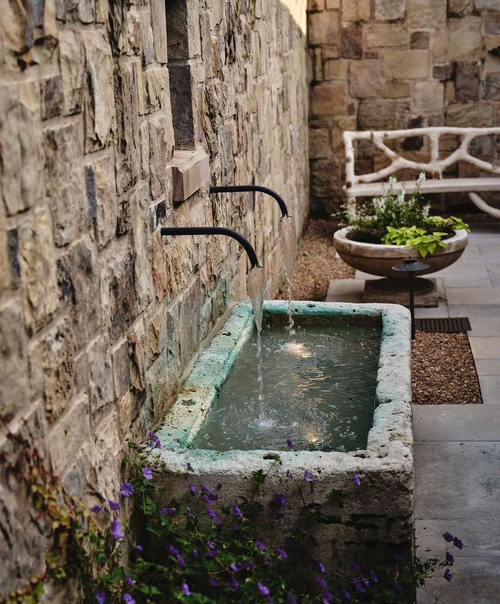 Outdoor stone fountain with water flowing from metal pipes into a basin, surrounded by purple flowers and stone wall. Outdoor bench visible.