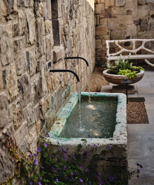 Outdoor stone fountain with water flowing from metal pipes into a basin, surrounded by purple flowers and stone wall. Outdoor bench visible.