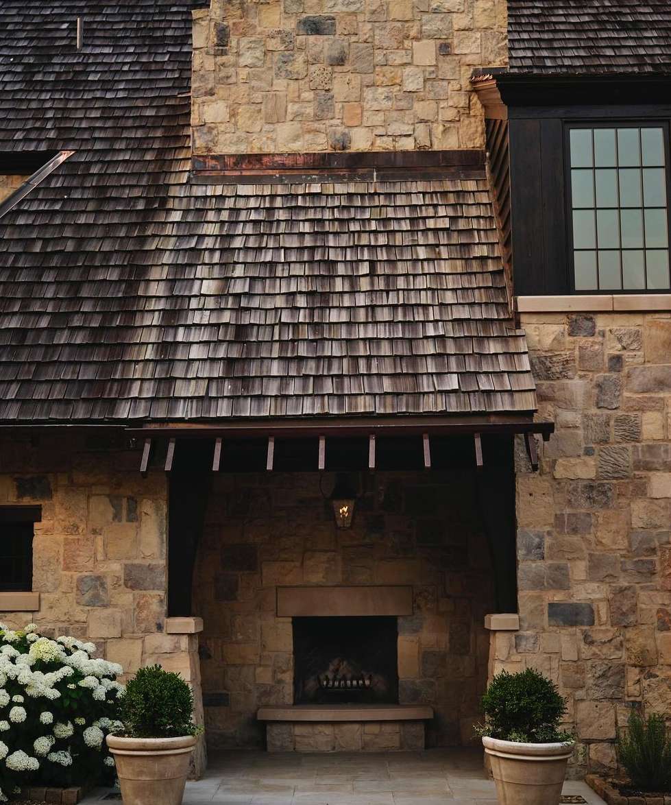 Exterior view of a stone house with a shingled roof, fireplace, and manicured greenery, showcasing architectural details and home design.