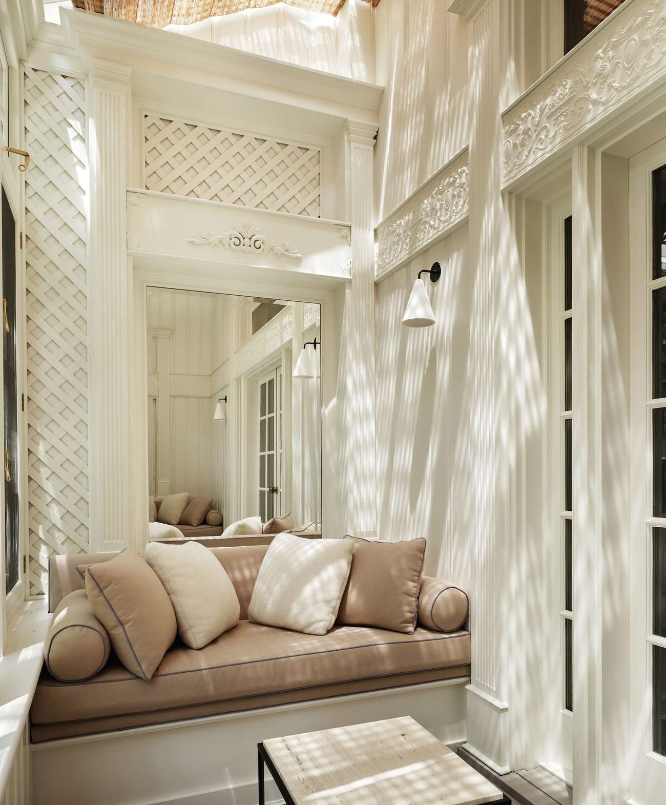 Indoor seating area with a tan cushioned bench, decorative pillows, and a marble-topped coffee table in a sun-drenched, white-walled room.