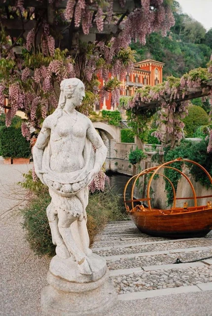 Stone statue of a woman stands in a lush garden with wisteria blooms, overlooking a boat and an ornate red building in the background.