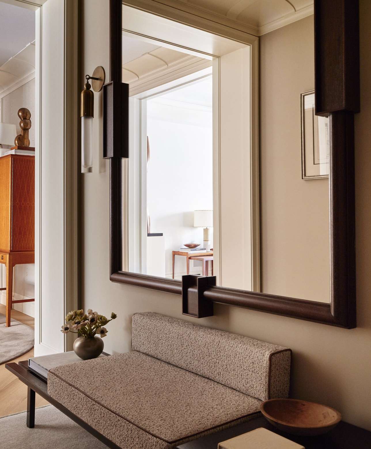 Interior with a large mirror, upholstered bench, and wooden bowl. Reflected light, neutral colors, and minimal decoration.