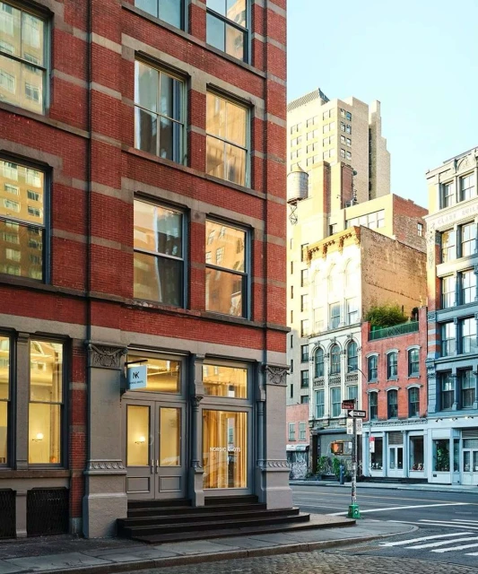 Exterior daytime view of a red brick building with large windows, grey doors, and steps on a sunny city street in New York.