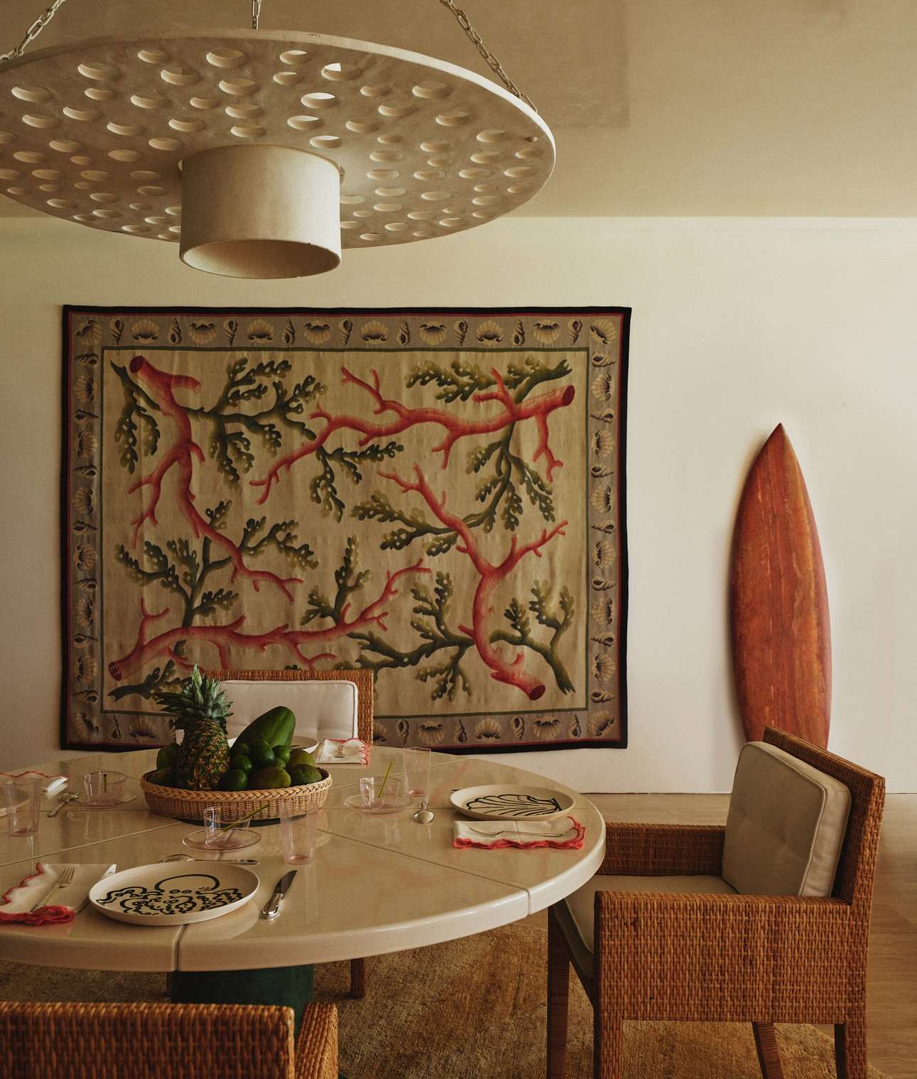 An indoor dining area features a round table set for a meal, surrounded by wicker chairs. A decorative rug and surfboard lean against the wall.