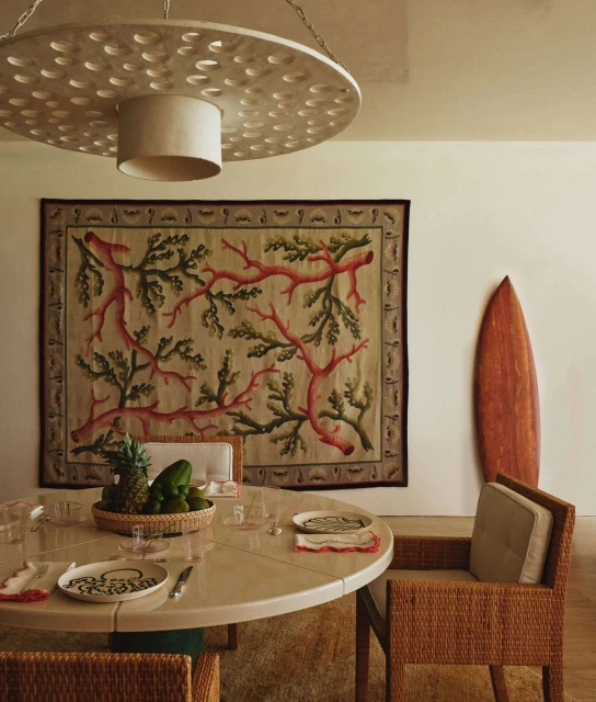An indoor dining area features a round table set for a meal, surrounded by wicker chairs. A decorative rug and surfboard lean against the wall.