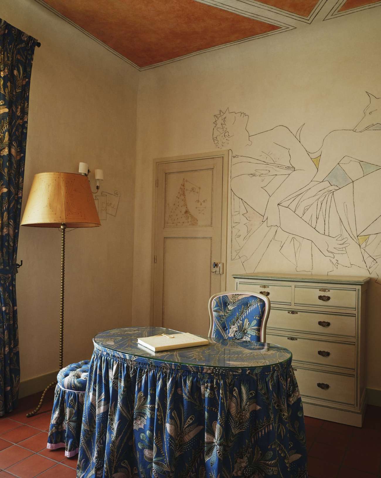 Indoor home study featuring a blue floral table and stool, ornate lamp, white dresser, and art on the walls, creating an elegant ambiance.