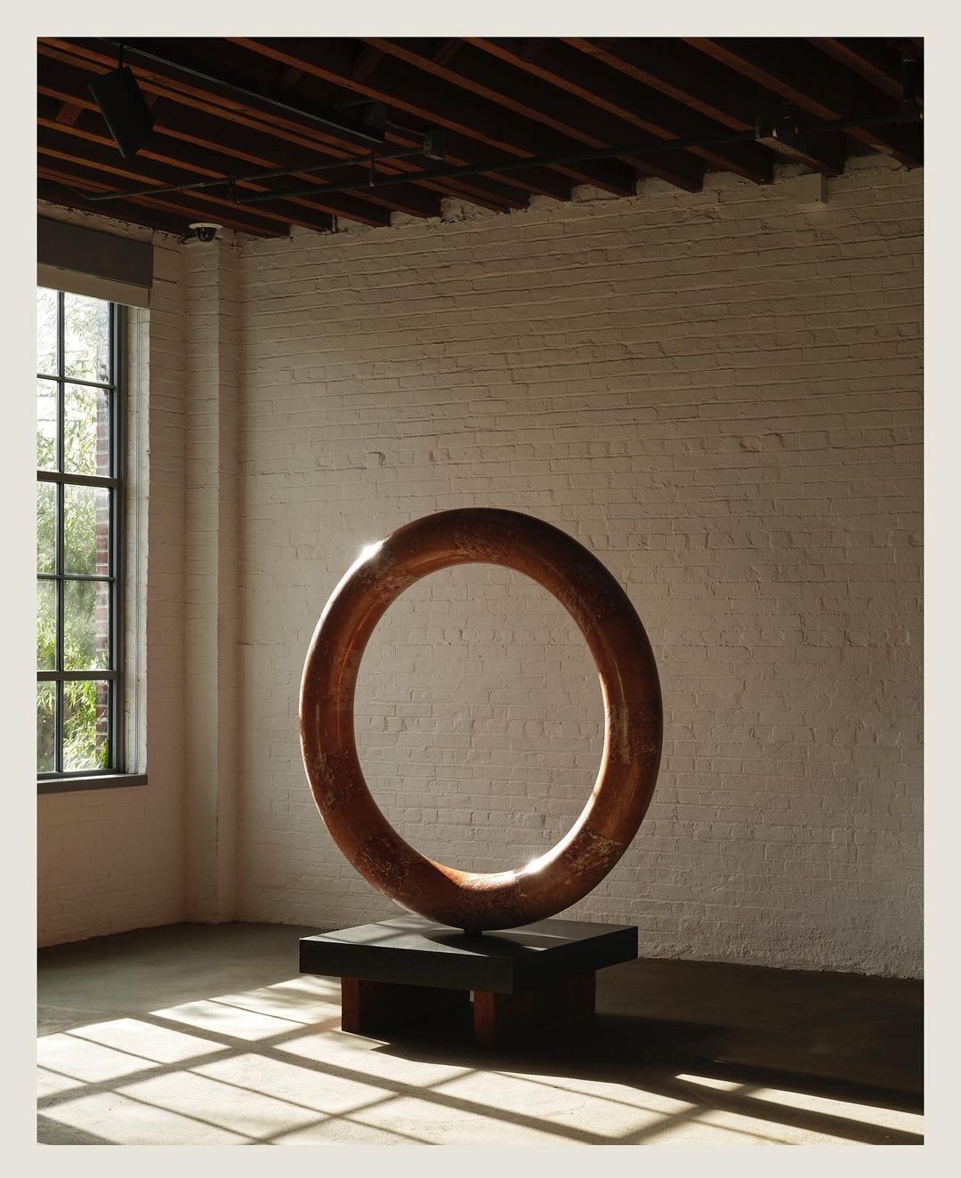 Modern art piece featuring a large circular sculpture made of copper, set against a brick wall and industrial-style flooring.