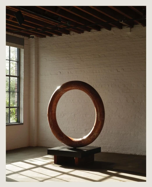 Modern art piece featuring a large circular sculpture made of copper, set against a brick wall and industrial-style flooring.