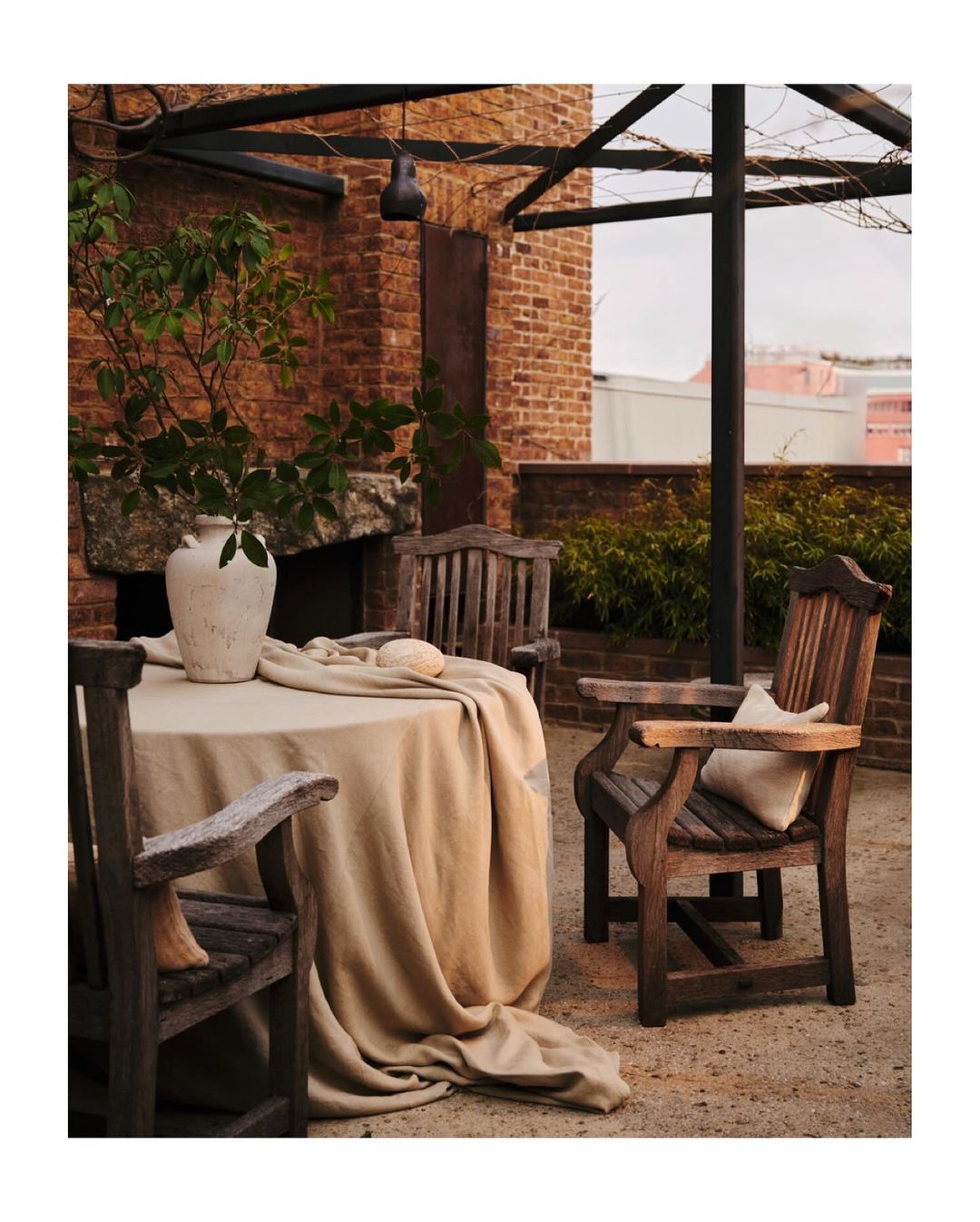 Outdoor dining setting featuring a beige-covered table, wooden chairs, a stone vase, and greenery, all set against a brick backdrop.