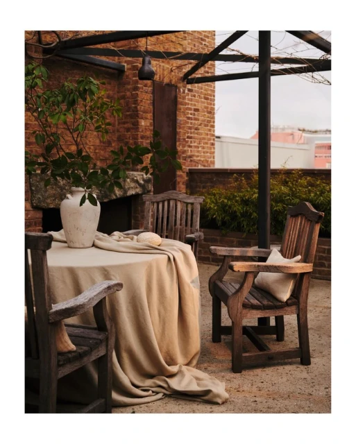 Outdoor dining setting featuring a beige-covered table, wooden chairs, a stone vase, and greenery, all set against a brick backdrop.