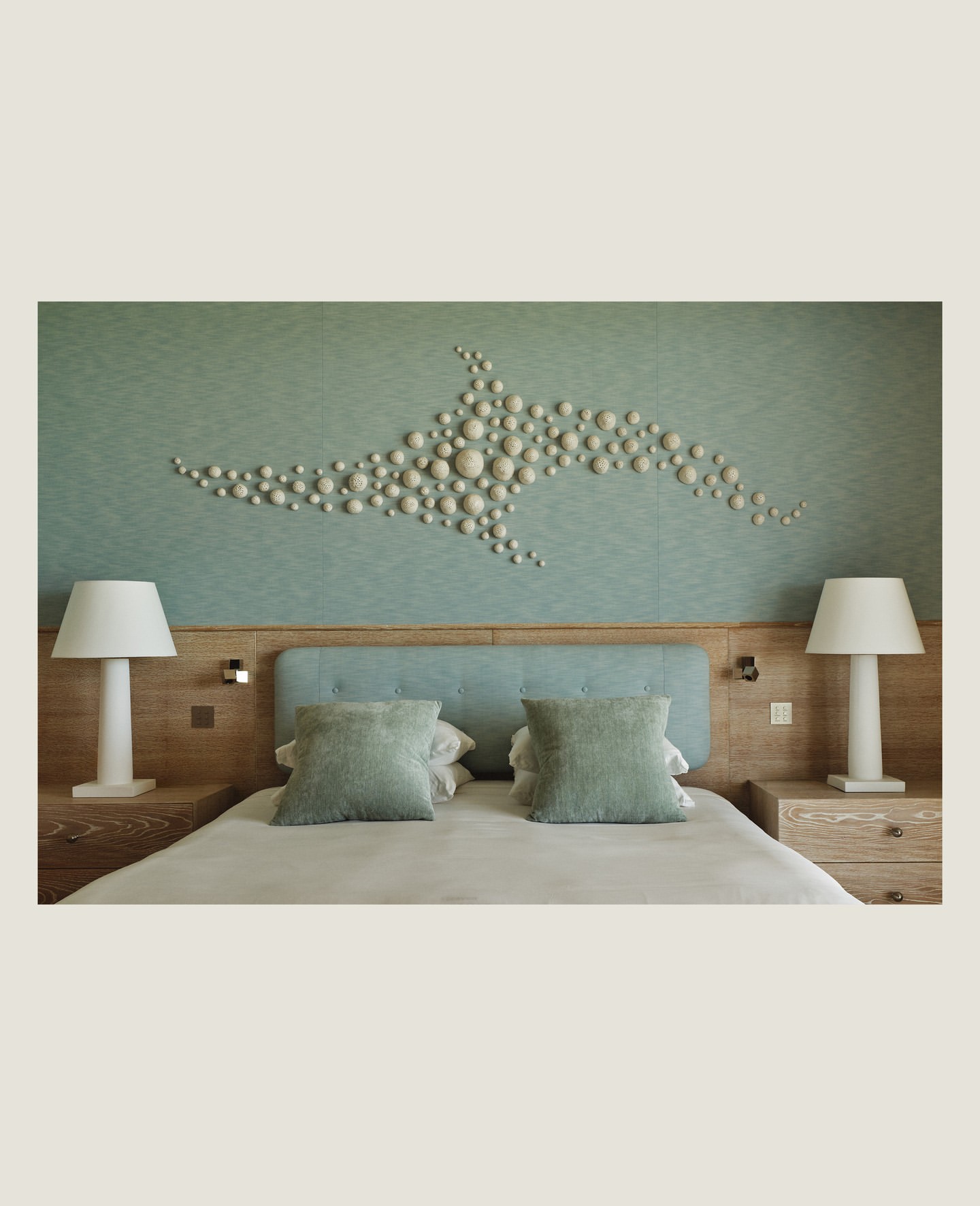 Hotel room interior featuring a large bed with green pillows, bedside tables with lamps, and an artistic wall decoration.