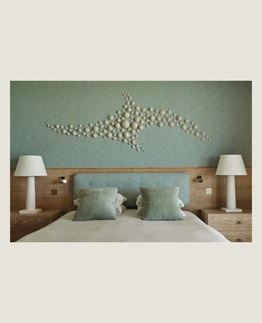 Hotel room interior featuring a large bed with green pillows, bedside tables with lamps, and an artistic wall decoration.