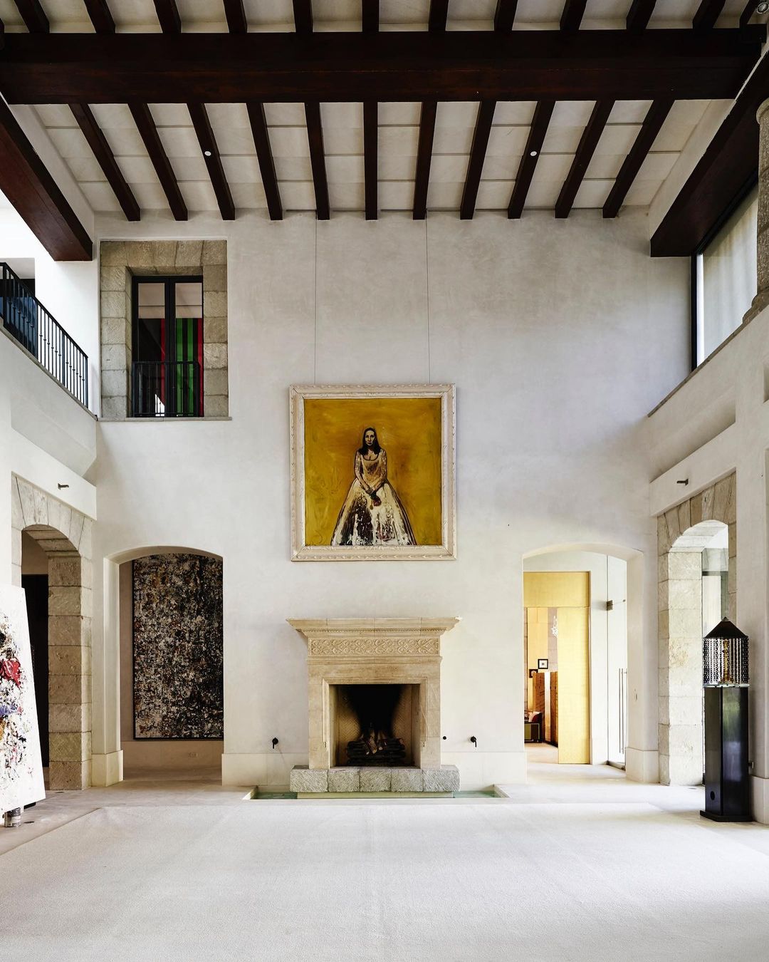 Interior view of a grand hall with a stone fireplace, an ornate painting, and a high ceiling with exposed wooden beams.