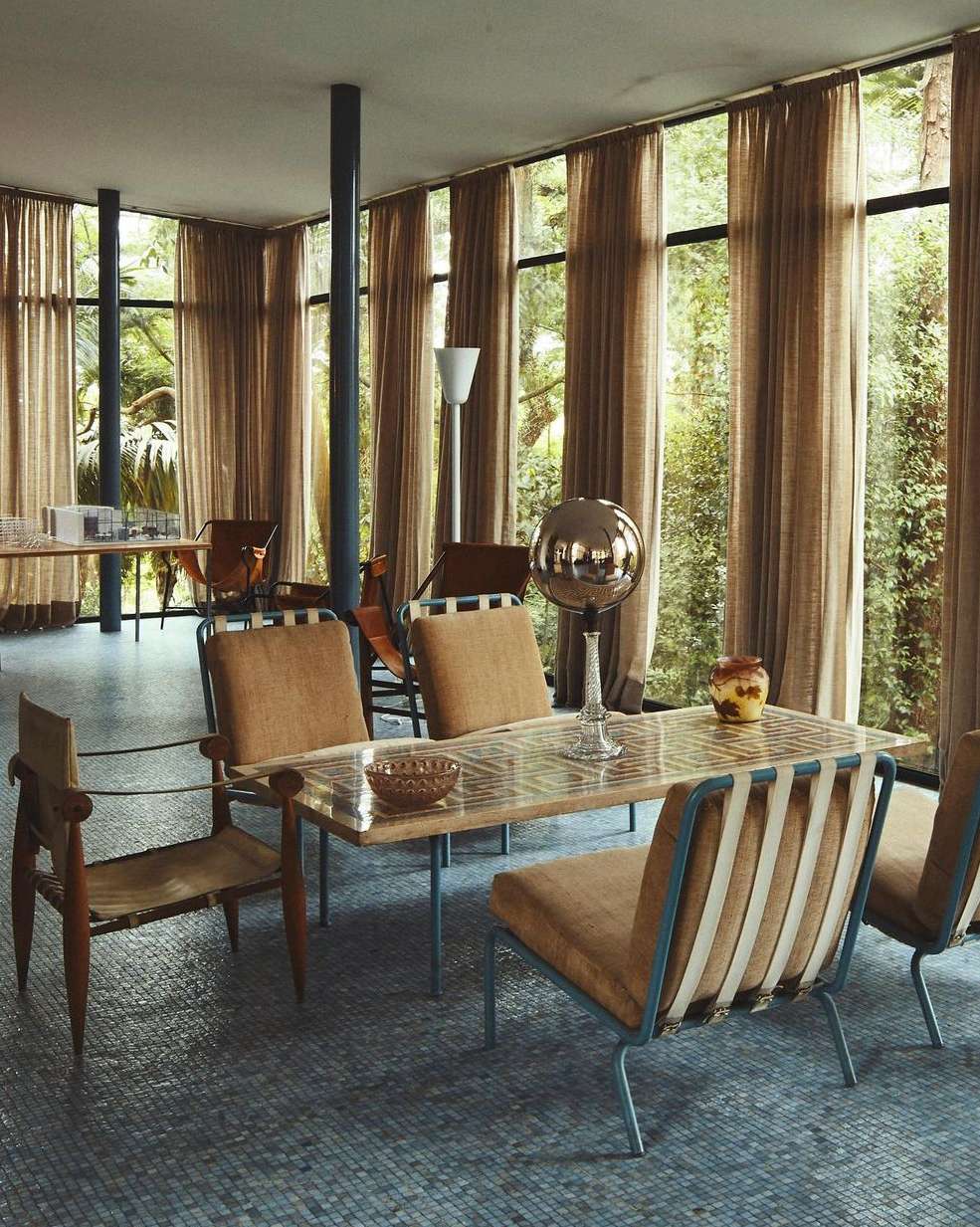 Modern dining room interior featuring a glass-topped table surrounded by mid-century modern chairs, with floor-to-ceiling windows overlooking greenery.