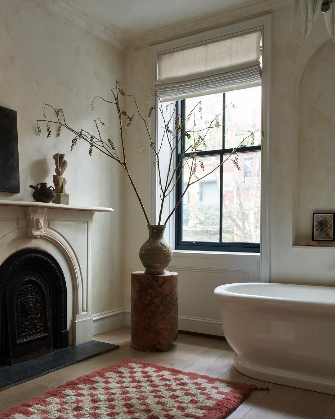 A bright bathroom showcases a white bathtub, fireplace, and a large vase with foliage on a red and white patterned rug.